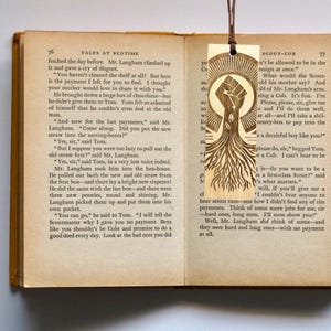 May include: A wooden bookmark with a raised fist and roots growing from the fist. The bookmark is brown and has a light brown wood grain. The text "Scout-Cub" is visible on the bookmark.