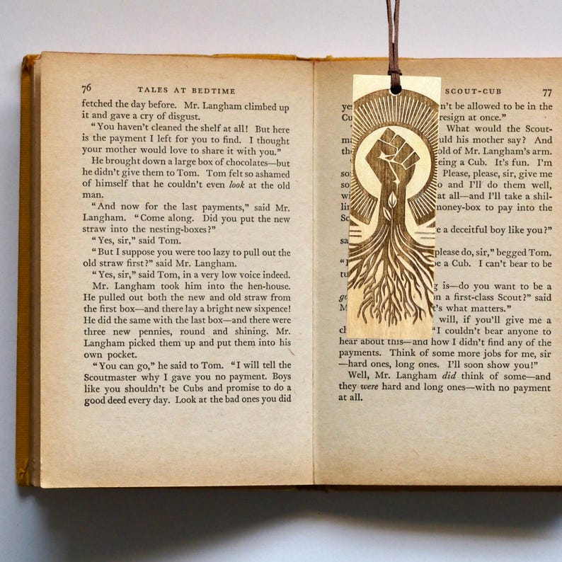 Handcrafted wooden bookmark featuring a laser-cut Black Pride fist with tree roots design, symbolizing strength, resilience, and heritage. Measuring 2”x6”, it includes a leatherette tassel and can be personalized with a quote or name on the back.