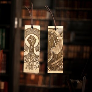 May include: Two wooden bookmarks with laser-engraved designs. One bookmark features a raised fist with roots growing from it, and the other features a stylized female figure with flowing hair.