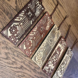 May include: Five wooden bookmarks with different designs. The bookmarks are laser cut and feature intricate patterns of gears, circuits, and floral designs. The bookmarks are all brown and beige in color and have a string attached to the top.