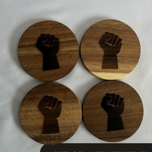 May include: Set of four wooden coasters with a dark brown finish. Each coaster features a black fist graphic. The coasters are round and have a natural wood grain pattern. The text "SET OF 4 STATEMENT COASTERS" is at the bottom.
