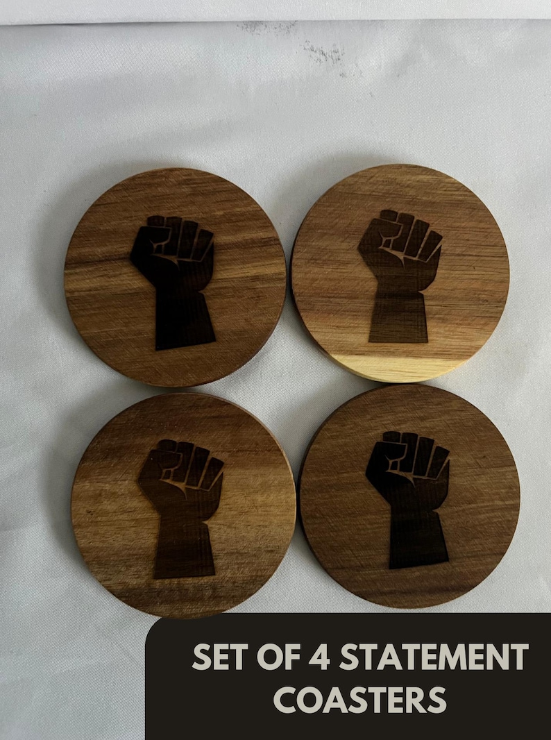 May include: Set of four wooden coasters with a dark brown finish. Each coaster features a black fist graphic. The coasters are round and have a natural wood grain pattern. The text "SET OF 4 STATEMENT COASTERS" is at the bottom.