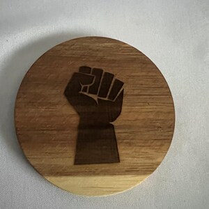 May include: A round wooden coaster with a dark brown fist graphic. The coaster is made of rich acacia grain and has the text "RICH ACACIA GRAIN" at the bottom. The coaster is a light brown color.