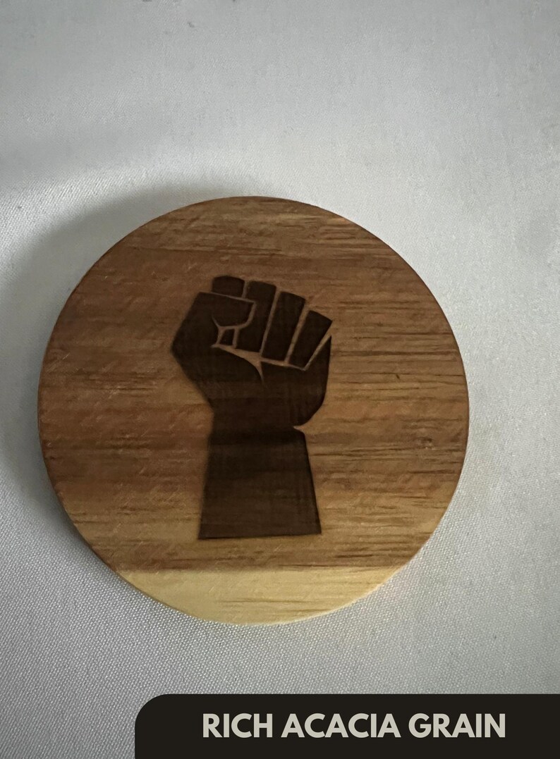 May include: A round wooden coaster with a dark brown fist graphic. The coaster is made of rich acacia grain and has the text "RICH ACACIA GRAIN" at the bottom. The coaster is a light brown color.