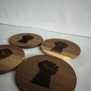 May include: Four wooden coasters with a dark brown finish and a black fist graphic. The coasters are round and feature a minimalist design. The text "MINIMALIST & MEANINGFUL" is at the bottom.