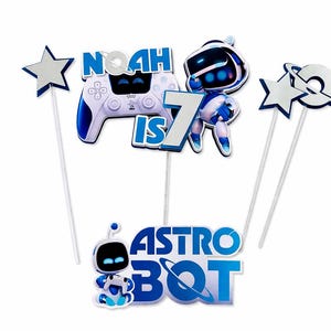 May include: A set of blue and white cake toppers featuring a video game controller, a robot character, stars, and planets. The text reads Noah is 7 and Astro Bot. Each cutout is attached to a thin white stick for placement on a cake or dessert.