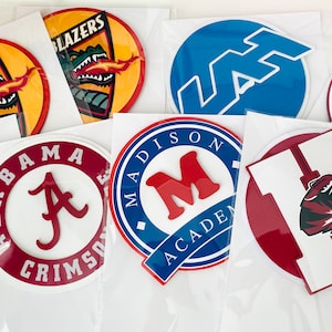 May include: A collection of nine round, colorful, iron-on patches with various school logos and mascots. The patches feature logos for UAB Blazers, Alabama Crimson Tide, Madison Academy, Lafayette, a tiger, and a tiger with the letter H.