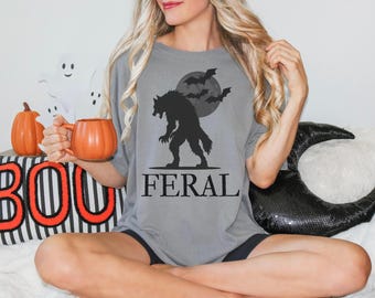 May include: A grey t-shirt featuring a black werewolf design, a full moon, and bats. The word "FERAL" is printed beneath the graphic. The image also includes a pumpkin-shaped mug and Halloween decorations.