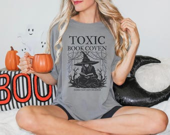 May include: Gray t-shirt with the text "TOXIC BOOK COVEN" and a graphic of a witch reading a book. The shirt is part of a Halloween theme, with a pumpkin-shaped mug and decorative pumpkins.