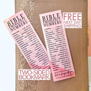 May include: A tan Bible with floral embossing, featuring two pink Bible Emergency Numbers bookmarks. The bookmarks list verses for various emotions and situations. The text "Two-Sided Bookmark" is visible.