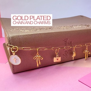 May include: A gold plated chain with multiple charms draped over a book. The charms include a white circular pendant with a cross, a bow, an envelope with a red heart, a small key, a second bow, and a round disc. Text reads GOLD PLATED CHAIN AND CHARMS.