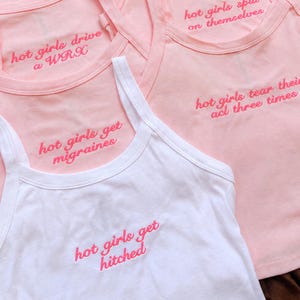May include: A collection of sleeveless tank tops in light pink and white, each featuring embroidered pink text. Phrases include hot girls spill on themselves, hot girls drive a WRX, hot girls tear their acl three times, hot girls get migraines, and hot girls get hitched.