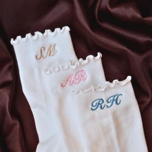 Personalized embroidered pilates socks with custom initials displayed on luxurious brown satin fabric featuring elegant white ribbed socks with feminine ruffle trim and soft pastel embroidery thread colors perfect for pilates lovers barre class outfits yoga girls bridesmaid gifts clean girl aesthetic fashion cozy lounge wear and trendy custom monogram accessories for women who love minimalist feminine styles and personalized boutique inspired gifts for birthdays holidays and bachelorette weekend