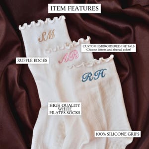 Custom monogram pilates socks photographed on rich satin fabric featuring delicate embroidered initials in soft pink blue and neutral thread colors on white ruffle socks designed for pilates class barre workouts yoga studio outfits and trendy clean girl aesthetic fashion perfect for bridesmaid proposal boxes bachelorette party gifts matching friend accessories travel essentials cozy self care gift baskets and personalized gifts for women who love elegant minimalist embroidered accessories
