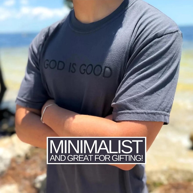May include: A dark gray t-shirt with the phrase "GOD IS GOOD" printed across the chest in black. The shirt has short sleeves and a crew neck. The text "MINIMALIST AND GREAT FOR GIFTING!" is in a white rectangle at the bottom.