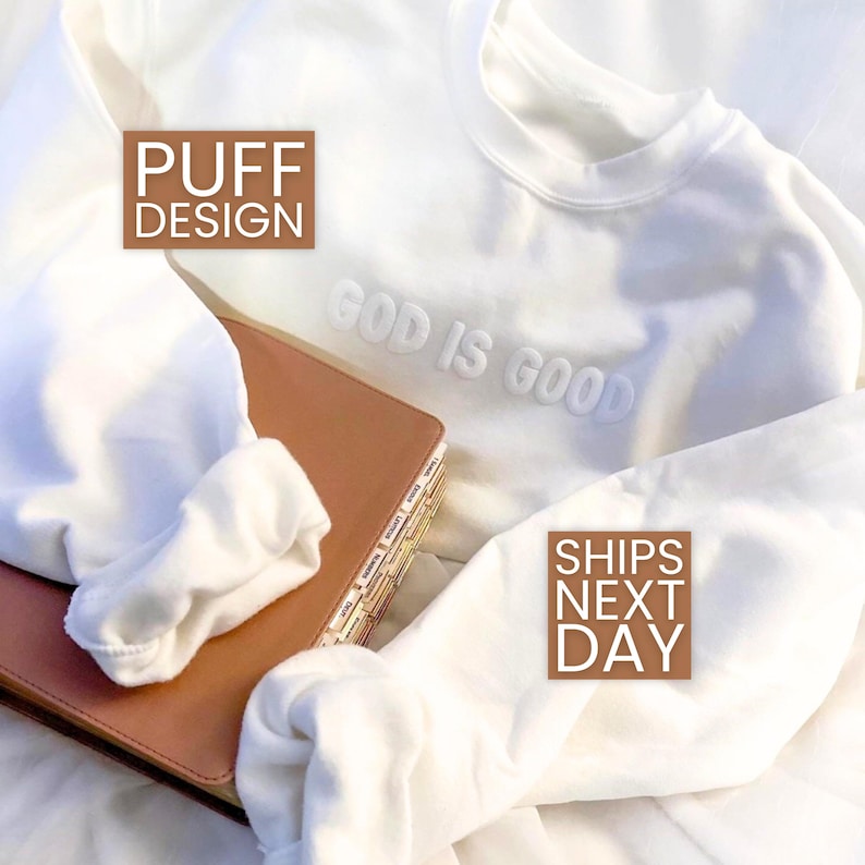 Puede incluir: Una sudadera blanca con la frase "GOD IS GOOD" en letras en relieve. La sudadera tiene un dise&ntilde;o abullonado y est&aacute; doblada sobre un libro marr&oacute;n. La imagen tambi&eacute;n incluye el texto "PUFF DESIGN".