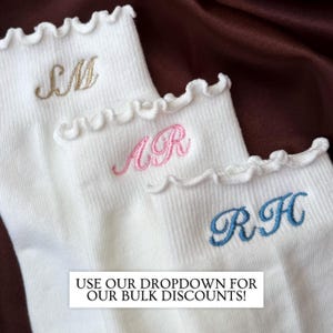 Embroidered initial socks with feminine ruffle edges styled on dark brown satin showcasing luxury inspired personalized pilates socks with elegant monogram embroidery ideal for barre class outfits yoga lovers bridesmaid gifts birthday gifts for women bachelorette weekend welcome bags and matching girls trip accessories featuring soft ribbed white socks with trendy clean aesthetic embroidery that creates a chic boutique style gift for everyday wear lounging and fitness fashion
