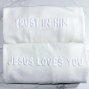 Puede incluir: Dos toallas blancas con texto en relieve. La toalla superior dice "TRUST IN HIM" y la toalla inferior dice "JESUS LOVES YOU". Las toallas est&aacute;n dobladas y apiladas sobre una superficie de m&aacute;rmol.