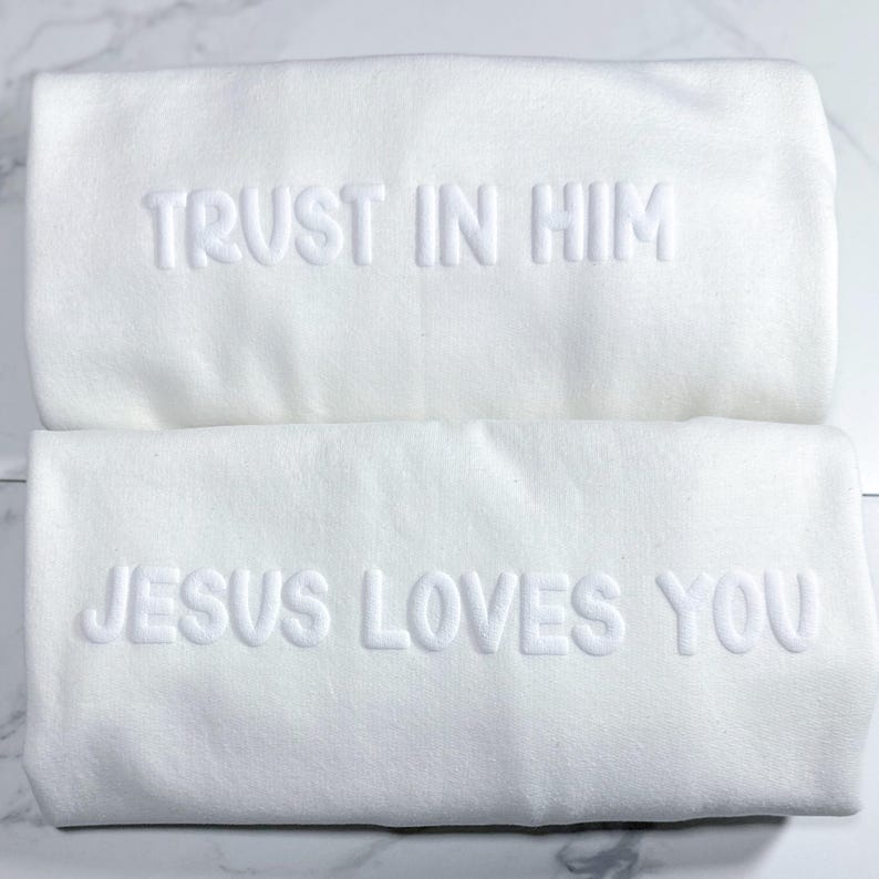 Puede incluir: Dos toallas blancas con texto en relieve. La toalla superior dice "TRUST IN HIM" y la toalla inferior dice "JESUS LOVES YOU". Las toallas est&aacute;n dobladas y apiladas sobre una superficie de m&aacute;rmol.