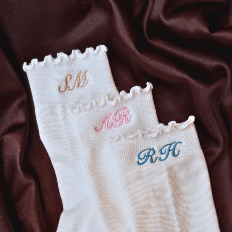 Personalized embroidered pilates socks with custom initials displayed on luxurious brown satin fabric featuring elegant white ribbed socks with feminine ruffle trim and soft pastel embroidery thread colors perfect for pilates lovers barre class outfits yoga girls bridesmaid gifts clean girl aesthetic fashion cozy lounge wear and trendy custom monogram accessories for women who love minimalist feminine styles and personalized boutique inspired gifts for birthdays holidays and bachelorette weekend