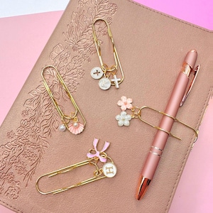 Può includere: Quattro fermagli in metallo dorato con ciondoli decorativi. Un fermaglio sostiene una conchiglia rosa e una perla, un altro una croce e la lettera K, uno un fiocco rosa e la lettera H, e uno una penna con due fiori. Il tutto su un taccuino color oro rosa.