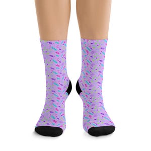 May include: A pair of crew-length socks featuring a light purple base color with a repeating pattern of small, colorful shapes including teal zigzags, pink squiggles, and dark purple dots. The socks have solid black toe and heel sections.