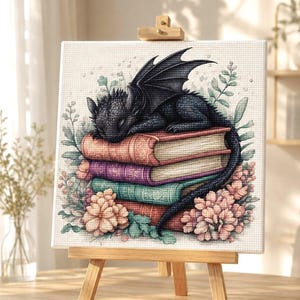 Puede incluir: Un lienzo cuadrado sobre un caballete de madera con una ilustración en punto de cruz de un pequeño dragón negro durmiendo sobre una pila de tres libros. Los libros son de tonos rosa, morado y turquesa, rodeados de flores de color rosa.