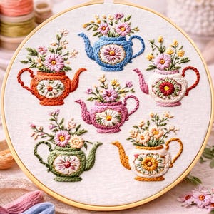 Puede incluir: Un bastidor de bordado circular de madera que contiene tela blanca con seis teteras bordadas a mano en azul, naranja, rosa, blanco, verde y amarillo. Cada tetera presenta un motivo floral central y est&aacute; llena de varias flores bordadas.