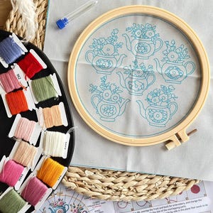 Puede incluir: Un kit de bordado que incluye una tela beige claro con cinco dise&ntilde;os azules de teteras y flores, tensada en un bastidor de pl&aacute;stico. Contiene un estuche transparente para agujas y un organizador negro con doce bobinas de hilo de colores.
