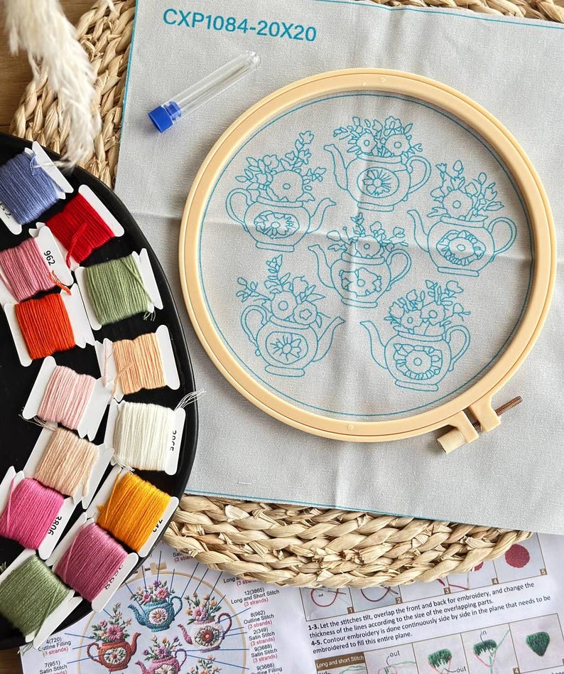 Puede incluir: Un kit de bordado que incluye una tela beige claro con cinco dise&ntilde;os azules de teteras y flores, tensada en un bastidor de pl&aacute;stico. Contiene un estuche transparente para agujas y un organizador negro con doce bobinas de hilo de colores.