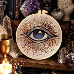 May include: Embroidered eye art in a wooden hoop. The eye is blue with gold accents, surrounded by gold rays. The background is a light beige fabric. The artwork is displayed on a wooden shelf with crystals, books, and candles.