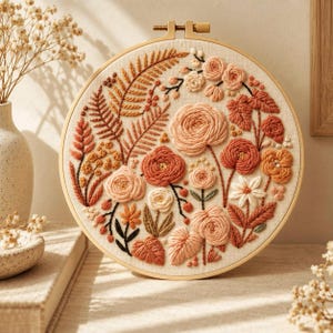 May include: A circular wooden embroidery hoop containing a piece of off-white fabric with a hand-stitched floral design. The embroidery features various flowers, leaves, and ferns in shades of peach, terracotta, rust, and cream thread.