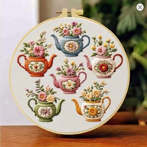 Puede incluir: Un bastidor de bordado circular de madera que contiene un panel de tela blanca con seis teteras bordadas a mano en azul, naranja, rosa, blanco, verde y amarillo. Cada tetera presenta un motivo central de margarita y arreglos florales.