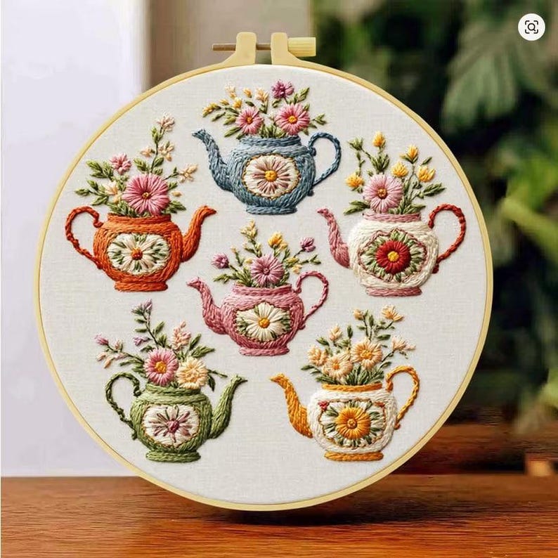Puede incluir: Un bastidor de bordado circular de madera que contiene un panel de tela blanca con seis teteras bordadas a mano en azul, naranja, rosa, blanco, verde y amarillo. Cada tetera presenta un motivo central de margarita y arreglos florales.