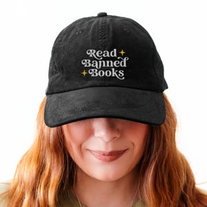 May include: Black corduroy baseball cap with the text "Read Banned Books" in white script. The hat has a curved brim and a structured crown. The cap is a classic style, suitable for everyday wear.