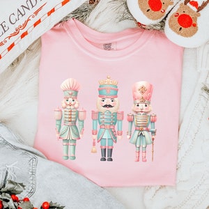 May include: A pink t-shirt with a graphic of three pastel-colored nutcracker figurines. The nutcrackers are wearing blue and gold uniforms and have pink accents. The shirt is laid out on a white fuzzy surface with a white knitted blanket and a red and white striped pillow.