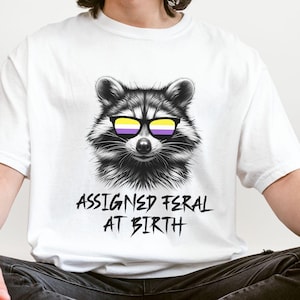 May include: White t-shirt featuring a black and white illustration of a raccoon wearing sunglasses with the colours of the non-binary pride flag. The text "ASSIGNED FERAL AT BIRTH" is printed below the raccoon.