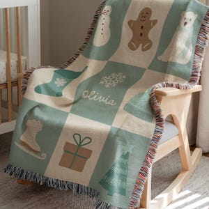 May include: A woven Christmas blanket with a checkerboard design, featuring festive illustrations of a snowman, gingerbread man, polar bear, stocking, snowflake, hat, ice skate, gift, and Christmas tree. The name "Olivia" is printed on the blanket, draped over a wooden rocking chair.
