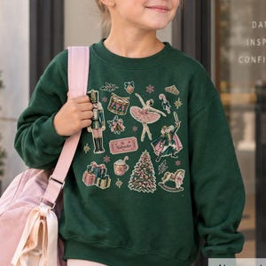 May include: A dark green crewneck sweatshirt featuring a festive Nutcracker-themed graphic print. The design includes a nutcracker soldier, a ballerina, a Christmas tree, wrapped gifts, a drum, a candy cane, and a ticket labeled The Nutcracker.