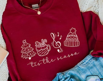 Op de afbeelding: Een rood sweatshirt met een wit grafisch ontwerp met een kerstboom, een mok warme chocolademelk, een muzieknoot en een wintermuts. De tekst "'tis the season" staat onder de afbeelding.