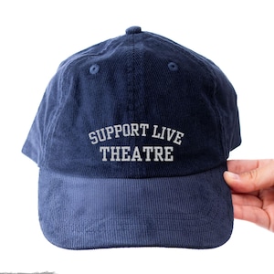 May include: Navy blue corduroy baseball cap with the text "SUPPORT LIVE THEATRE" in white block letters. The cap has a curved brim and a rounded top. The cap is held by a hand.