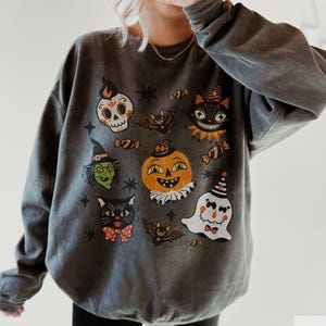 May include: A charcoal grey long-sleeve sweatshirt featuring a vintage-style Halloween graphic print. The design includes a skull, a witch, a jack-o'-lantern, a ghost, black cats, bats, and candy icons in orange, white, and black. The text Pepper is visible.