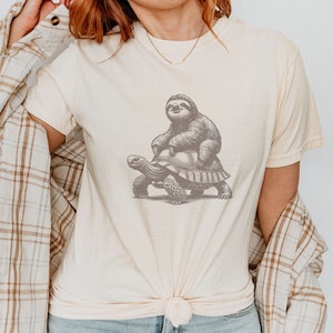 May include: A cream-colored t-shirt with a graphic of a sloth riding a turtle. The sloth and turtle are in a dark gray color.
