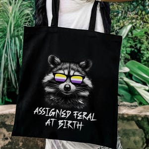 May include: Black tote bag featuring a raccoon wearing non-binary pride flag sunglasses. The raccoon is in black and white. The text "ASSIGNED FERAL AT BIRTH" is written in white below the raccoon.