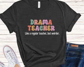May include: A dark gray t-shirt with the text "DRAMA TEACHER" in colorful letters. The text below reads "Like a regular teacher, but weirder."