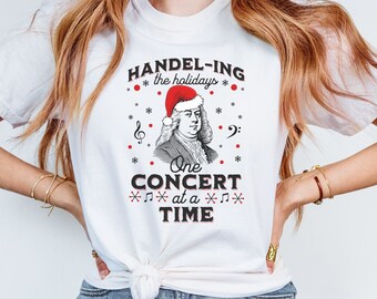May include: White t-shirt with a graphic of a man wearing a Santa hat and the text "HANDEL-ING the holidays One CONCERT at a TIME".