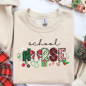 May include: A beige sweatshirt with the words "school nurse" in green, red, and black letters. The letters are decorated with Christmas-themed designs, including a bow, baubles, and a stethoscope.