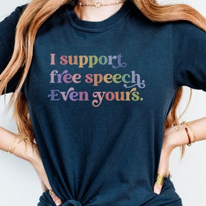 May include: Navy blue t-shirt with the text "I support free speech. Even yours." in rainbow colours. The shirt is knotted at the waist. The shirt is a casual wear item.