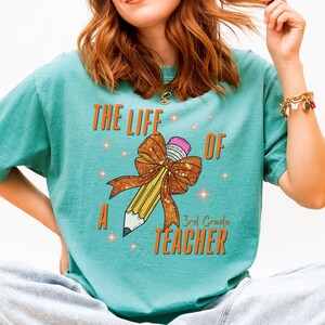 Peut inclure: T-shirt de couleur sarcelle avec l'inscription "THE LIFE OF A 3rd GRADE TEACHER" en orange. Un grand crayon avec un nœud est au centre. Le crayon est jaune avec une gomme rose et une pointe blanche. Le t-shirt est de style décontracté.