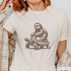 May include: An ivory t-shirt featuring a dark brown graphic illustration of a sloth riding on the back of a tortoise. The shirt is worn with a tan and white plaid button-down shirt layered over it, and the word Ivory is printed in the corner.